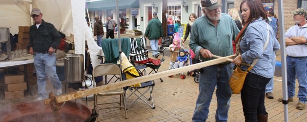 Visitors gathered at an outdoor event, with tents and stalls set up around them. An elderly man and a young woman work together, using a long wooden tool to stir something in a large pot. Other people watch them, and the scene has a lively, festive atmosphere.