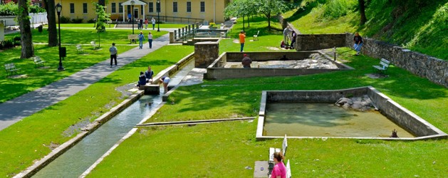 A scenic park with green lawns and small stream-fed pools. People are strolling and sitting on benches, enjoying the sunny day. Stone pathways lead through the park with a historic building and trees in the background. Ducks swim in the ponds.