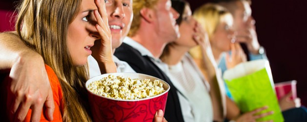 A group of people in a movie theater are watching a film. The person in the foreground, holding a red bucket of popcorn, covers their face with a hand, appearing scared. Others in the row are also reacting with various expressions, holding popcorn.