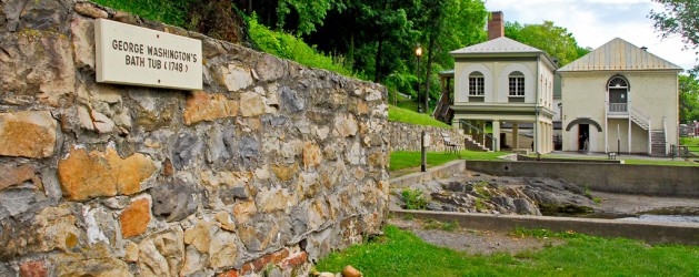 A historic stone wall with a sign reading "George Washington's Bath Tub (1748)" in front of a building with arched windows. Lush greenery surrounds the area, and the scene appears to be part of a historical site.