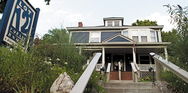 Wide stone stairs with wooden railings lead up to the entrance of a two-story blue house with white trim and a wrap-around porch. The property is surrounded by greenery and a sign to the left reads, "Lot 12 Public House.