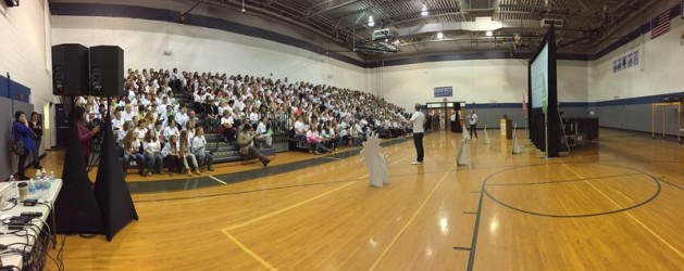 A large group of people sit on bleachers inside a gymnasium, attentively watching a person speaking on stage. The gym has high ceilings with visible lighting fixtures, and there are several screens set up for presentations.