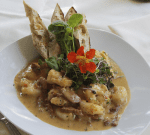 A white plate contains a dish of creamy potato gnocchi with pieces of meat in a sauce, garnished with microgreens, an orange edible flower, and accompanied by slices of toasted bread. The background shows a white tablecloth and a folded napkin.