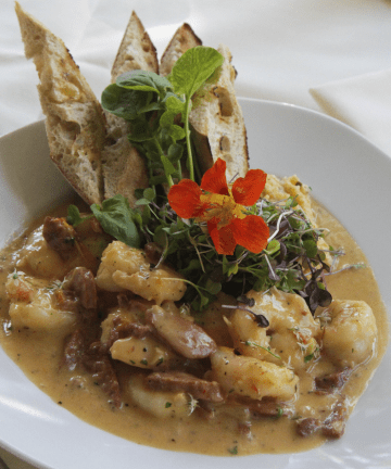 A white plate contains a dish of creamy potato gnocchi with pieces of meat in a sauce, garnished with microgreens, an orange edible flower, and accompanied by slices of toasted bread. The background shows a white tablecloth and a folded napkin.