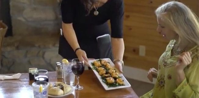 A server in a black dress places a rectangular plate of food on a table in front of a seated woman with long blonde hair wearing a green blouse. The table has glasses of water, wine, and additional food items.