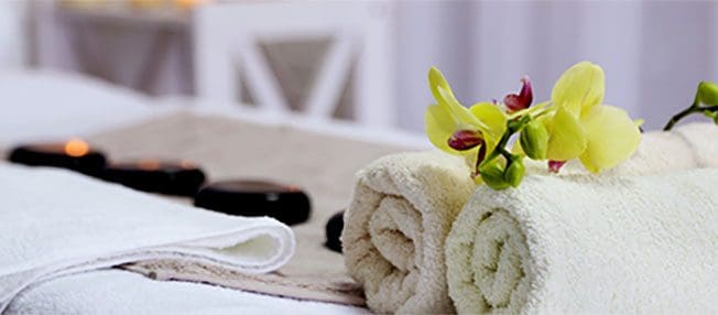 Rolled white towels decorated with a yellow orchid flower are placed on a massage table in a serene spa setting. In the background, black massage stones are neatly arranged, creating a relaxing and tranquil atmosphere.