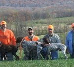 Four men wearing outdoor attire and bright orange caps pose with three dogs in a grassy area with a wooded backdrop. Two of the dogs are black-and-white, and the third is brown. The scene appears to be set in the countryside during autumn.