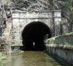 An old stone tunnel entrance with an arch shape, partially submerged in water. The tunnel is surrounded by rocky terrain and greenery. The water leads into the dark interior of the tunnel. A small ledge with a railing runs along the right side of the tunnel.