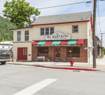 A two-story cream and red building with a sign reading "Mi Ranchito Mexican Restaurant." It features a storefront with large windows and flowerpots, a red fire hydrant on the corner, and utility poles along the sidewalk, set in a small town with greenery in the background.