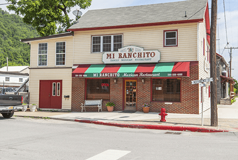 A two-story cream and red building with a sign reading "Mi Ranchito Mexican Restaurant." It features a storefront with large windows and flowerpots, a red fire hydrant on the corner, and utility poles along the sidewalk, set in a small town with greenery in the background.