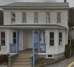 A two-story white building with a gray metal roof featuring a front porch supported by blue columns. A sign reading "Healing Touch Clinic" is displayed near the porch. Concrete steps and a wheelchair ramp lead up to the entrance. The building is located on a sloped street.