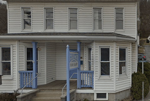 A two-story white building with a gray metal roof featuring a front porch supported by blue columns. A sign reading "Healing Touch Clinic" is displayed near the porch. Concrete steps and a wheelchair ramp lead up to the entrance. The building is located on a sloped street.