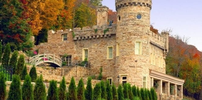 A picturesque stone castle with a turret and crenellated walls is nestled among vibrant autumn trees. A small arched bridge crosses a path by the entrance, and neatly trimmed bushes form a line in the foreground. The sky is a soft purple hue.