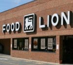 A brick building with a large "Food Lion" sign above the entrance. The white sign includes a lion logo in between the words "Food" and "Lion." The windows below the sign display various promotional posters.