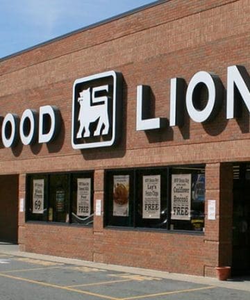 A brick building with a large "Food Lion" sign above the entrance. The white sign includes a lion logo in between the words "Food" and "Lion." The windows below the sign display various promotional posters.