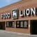 A brick building with a large "Food Lion" sign above the entrance. The white sign includes a lion logo in between the words "Food" and "Lion." The windows below the sign display various promotional posters.