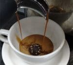 A coffee machine pours espresso into a white cup with a chocolate truffle inside. The cup sits on a matching saucer, and the espresso is beginning to fill the cup, partially melting the truffle.