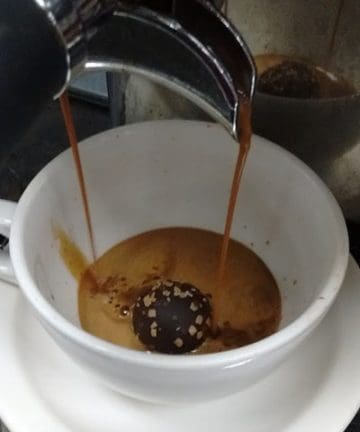 A coffee machine pours espresso into a white cup with a chocolate truffle inside. The cup sits on a matching saucer, and the espresso is beginning to fill the cup, partially melting the truffle.