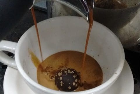 A coffee machine pours espresso into a white cup with a chocolate truffle inside. The cup sits on a matching saucer, and the espresso is beginning to fill the cup, partially melting the truffle.