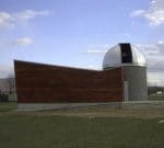 A small observatory building with a dome-shaped roof and a wooden exterior wall stands on a grassy area under a partly cloudy sky.