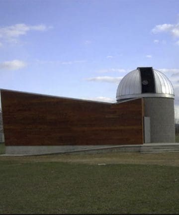 A small observatory building with a dome-shaped roof and a wooden exterior wall stands on a grassy area under a partly cloudy sky.