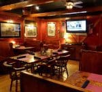 A cozy restaurant interior with wooden furniture and a ceiling fan. There are several tables set with folded napkins, a TV mounted on the wall showing a news channel, American flags, and framed pictures on red-painted walls, creating a warm and inviting atmosphere.