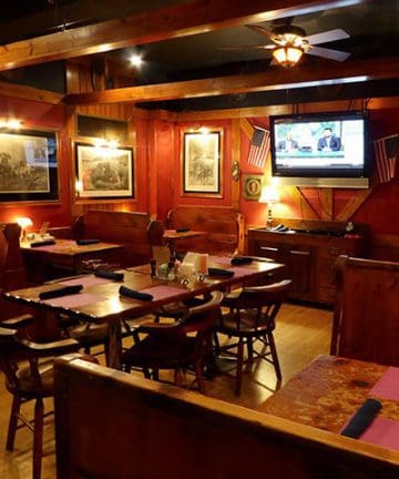 A cozy restaurant interior with wooden furniture and a ceiling fan. There are several tables set with folded napkins, a TV mounted on the wall showing a news channel, American flags, and framed pictures on red-painted walls, creating a warm and inviting atmosphere.