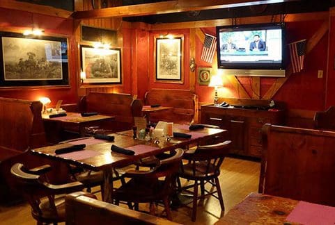 A cozy restaurant interior with wooden furniture and a ceiling fan. There are several tables set with folded napkins, a TV mounted on the wall showing a news channel, American flags, and framed pictures on red-painted walls, creating a warm and inviting atmosphere.