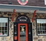 Entrance to a building with a stone facade and a sign above the door reading "Tasting Room." The door is red with a window displaying an image of an olive and text that says "The Naked Olive." The building has large windows and decorative trim.