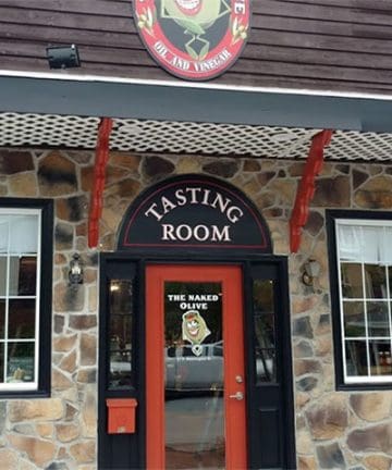 Entrance to a building with a stone facade and a sign above the door reading "Tasting Room." The door is red with a window displaying an image of an olive and text that says "The Naked Olive." The building has large windows and decorative trim.