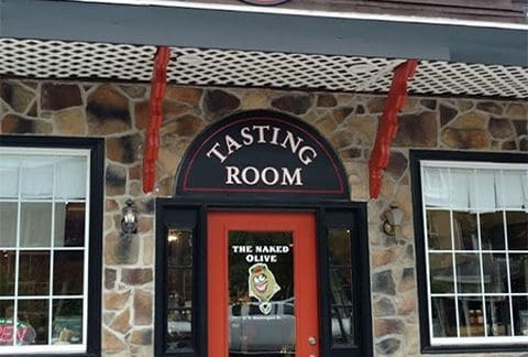 Entrance to a building with a stone facade and a sign above the door reading "Tasting Room." The door is red with a window displaying an image of an olive and text that says "The Naked Olive." The building has large windows and decorative trim.