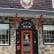 Entrance to a building with a stone facade and a sign above the door reading "Tasting Room." The door is red with a window displaying an image of an olive and text that says "The Naked Olive." The building has large windows and decorative trim.