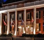 A large, illuminated brick building with white columns and a balcony adorned with lattice railings. Several flags hang from the building's front, and there are rocking chairs on the porch. White lights highlight the entrance, giving the building a welcoming look.
