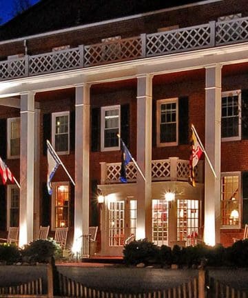 A large, illuminated brick building with white columns and a balcony adorned with lattice railings. Several flags hang from the building's front, and there are rocking chairs on the porch. White lights highlight the entrance, giving the building a welcoming look.