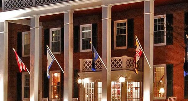 A large, illuminated brick building with white columns and a balcony adorned with lattice railings. Several flags hang from the building's front, and there are rocking chairs on the porch. White lights highlight the entrance, giving the building a welcoming look.