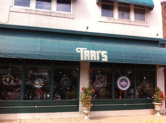 A storefront with a green awning displaying the name "Gary's" in white text. The facade has large windows that show various decorative items, including circular stained glass pieces. Two potted plants are placed on either side of the entrance.