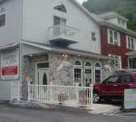 A two-story building with a stone facade on the lower level and white siding on the upper level. The building features arched windows and a small balcony. A red SUV is parked in front, and a sign reads "Maria's Garden & Inn Bed & Breakfast, Bakery, Restaurant.