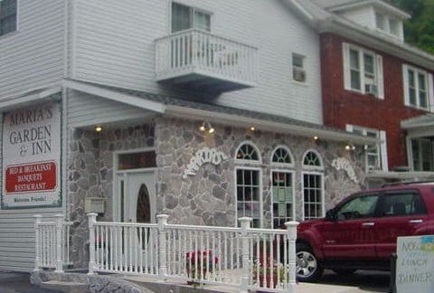A two-story building with a stone facade on the lower level and white siding on the upper level. The building features arched windows and a small balcony. A red SUV is parked in front, and a sign reads "Maria's Garden & Inn Bed & Breakfast, Bakery, Restaurant.