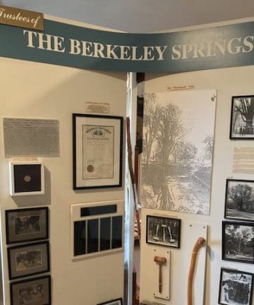 A display board titled "Trustees of The Berkeley Springs" showcases a collection of historical documents, framed photographs, and artifacts, including a gavel and a walking cane. The board is situated in a well-lit room.