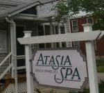 A storefront featuring a sign that reads "Atasia Spa, Berkeley Springs, W.V." The building is light gray with steps leading up to the entrance and a small overhang. There is also a whiteboard with additional information partially visible on the left side. Trees frame the scene.