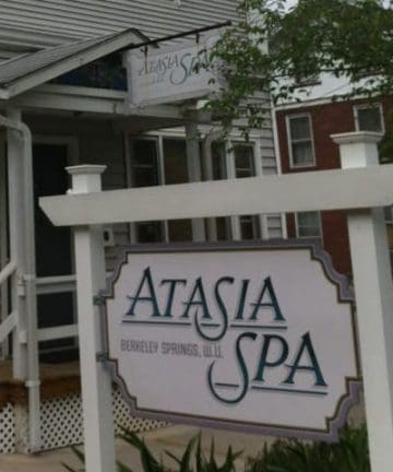 A storefront featuring a sign that reads "Atasia Spa, Berkeley Springs, W.V." The building is light gray with steps leading up to the entrance and a small overhang. There is also a whiteboard with additional information partially visible on the left side. Trees frame the scene.