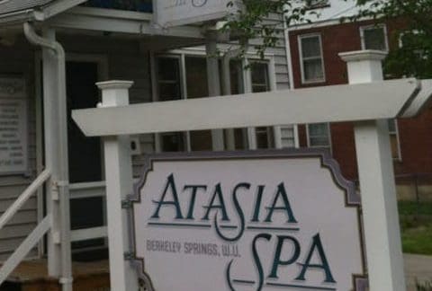 A storefront featuring a sign that reads "Atasia Spa, Berkeley Springs, W.V." The building is light gray with steps leading up to the entrance and a small overhang. There is also a whiteboard with additional information partially visible on the left side. Trees frame the scene.