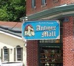 A brick building corner with a bright blue sign reading "Antique Mall." The sign features an image of a woman in profile, wearing a red hat and looking down. Below the sign, a window displays various items for sale inside the mall.