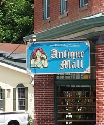 A brick building corner with a bright blue sign reading "Antique Mall." The sign features an image of a woman in profile, wearing a red hat and looking down. Below the sign, a window displays various items for sale inside the mall.