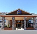 The image shows the front entrance of a Best Western hotel. The building features a wooden facade with stone pillars supporting a large overhang. The Best Western sign is prominently displayed above the entrance. An American flag is visible to the right of the entrance.