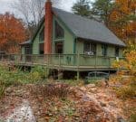 A cabin with a steep roof, green exterior, and prominent brick chimney rests in an autumnal setting. The cabin is surrounded by a wooden deck, with a pond filled with fallen leaves and water lilies in the foreground. Trees with fall foliage encircle the scene.
