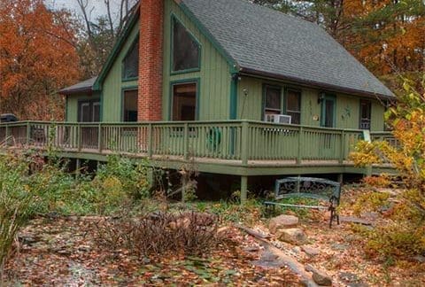 A cabin with a steep roof, green exterior, and prominent brick chimney rests in an autumnal setting. The cabin is surrounded by a wooden deck, with a pond filled with fallen leaves and water lilies in the foreground. Trees with fall foliage encircle the scene.