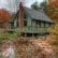 A cabin with a steep roof, green exterior, and prominent brick chimney rests in an autumnal setting. The cabin is surrounded by a wooden deck, with a pond filled with fallen leaves and water lilies in the foreground. Trees with fall foliage encircle the scene.