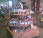 A retail store display featuring a variety of glassware, including jars, glasses, and cups arranged on a multi-tiered wooden table. The shelves around the display hold various other products, and the room is well lit with natural light from windows.