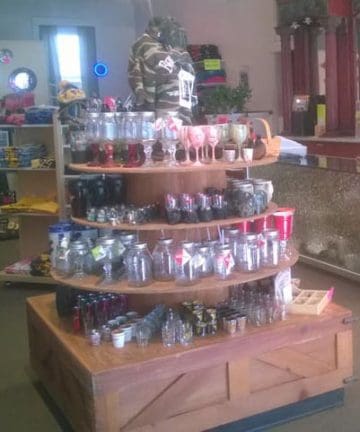 A retail store display featuring a variety of glassware, including jars, glasses, and cups arranged on a multi-tiered wooden table. The shelves around the display hold various other products, and the room is well lit with natural light from windows.